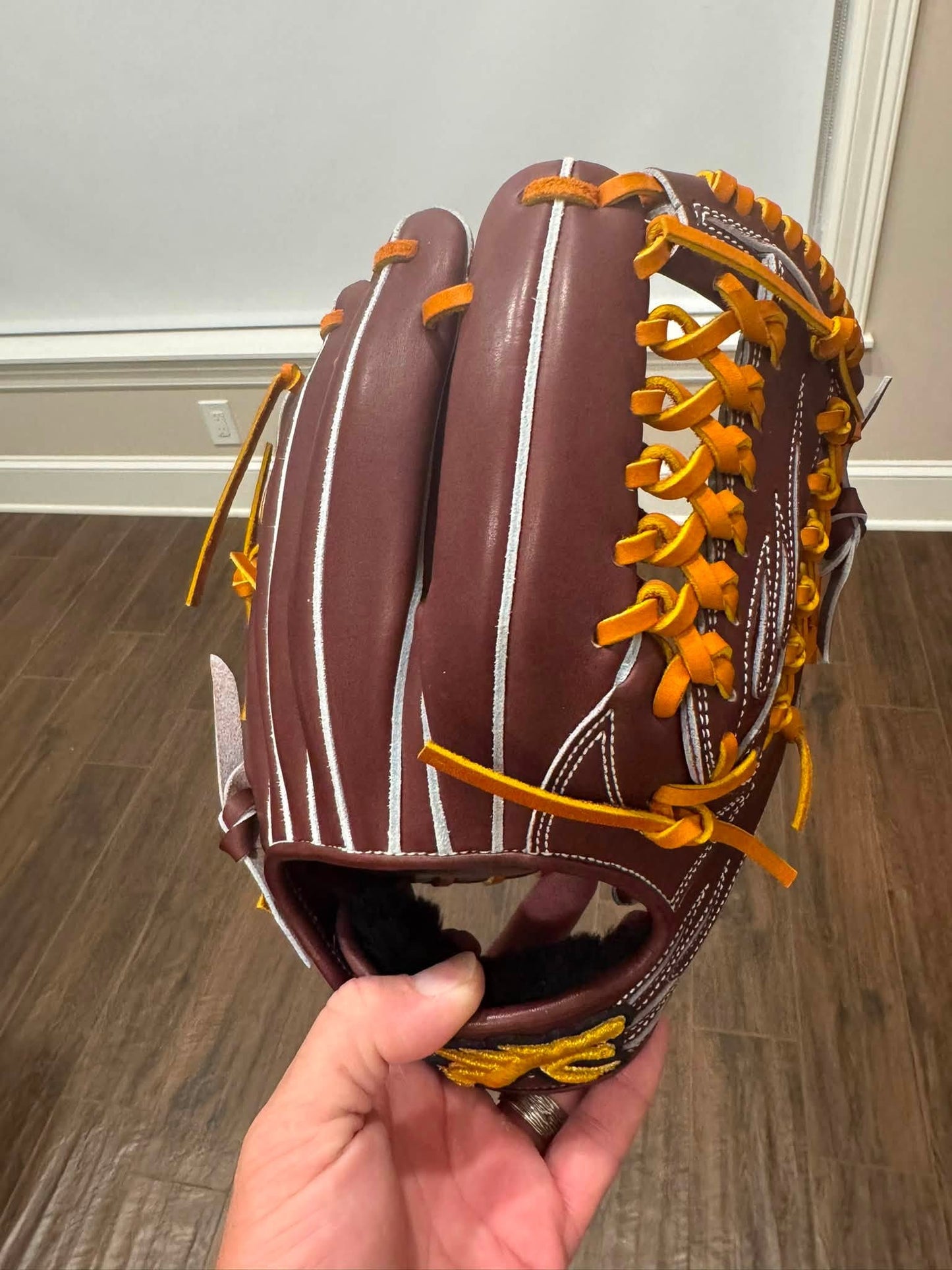 A brown leather baseball glove with orange laces and white pinstripes, held in a person's hand against an indoor background of wood flooring and a white wall.