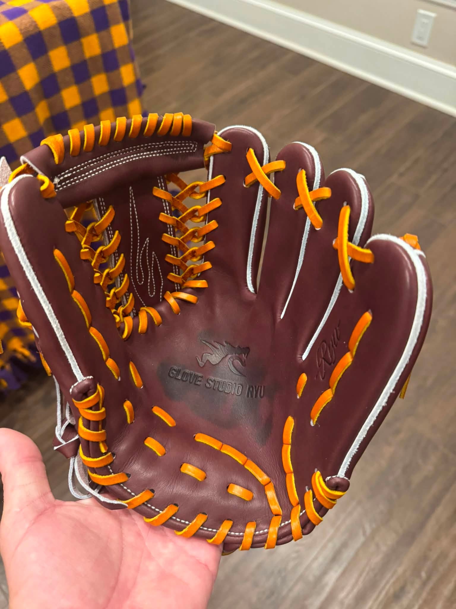 A brown leather baseball glove held in a hand, featuring bright orange laces and white piping, with a yellow and purple plaid blanket in the background.